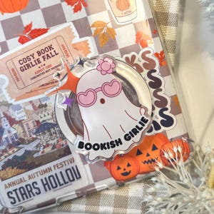 Puede incluir: Una funda transparente para teléfono con temática otoñal, con pegatinas de un fantasma, calabazas y hojas. La funda tiene las palabras "hello FALL" y "BOOKISH GIRLIE". El fondo es un patrón de cuadros.