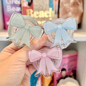 May include: Three clear acrylic bow-shaped phone grips in pastel colors: mint green, light blue, and pink. The grips are held in front of a bookshelf with books and a decorative item.