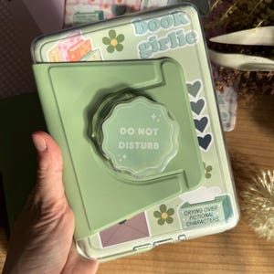 以下が含まれることがあります： 花柄とハートのステッカーで飾られた、ポップアップグリップ付きのライトグリーンの携帯電話ケース。「book girlie」と「Do Not Disturb」の文字がプリントされています。「Probably reading a romance book」という文字が入った2つ目のケースが見えます。