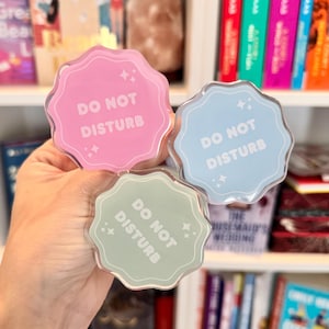 Puede incluir: Tres letreros acrílicos con bordes festoneados en rosa, azul y verde, cada uno con las palabras "DO NOT DISTURB" en blanco. Los letreros tienen un diseño de estrella y se sostienen frente a una estantería llena de libros.