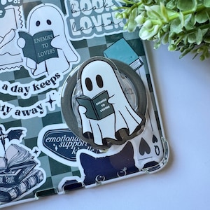 以下が含まれることがあります： 「ENEMIES TO LOVERS」の文字が入った本を読むお化けのイラストが描かれたクリアなスマホケース。本と蜂のイラストのステッカーも付いています。背景は緑と白のチェック柄です。