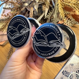 May include: Two black and clear phone grips with the text "ANTISOCIAL BOOK CLUB" and an open book graphic. The grips are round and held in front of a blurred background with dried flowers and a phone case.