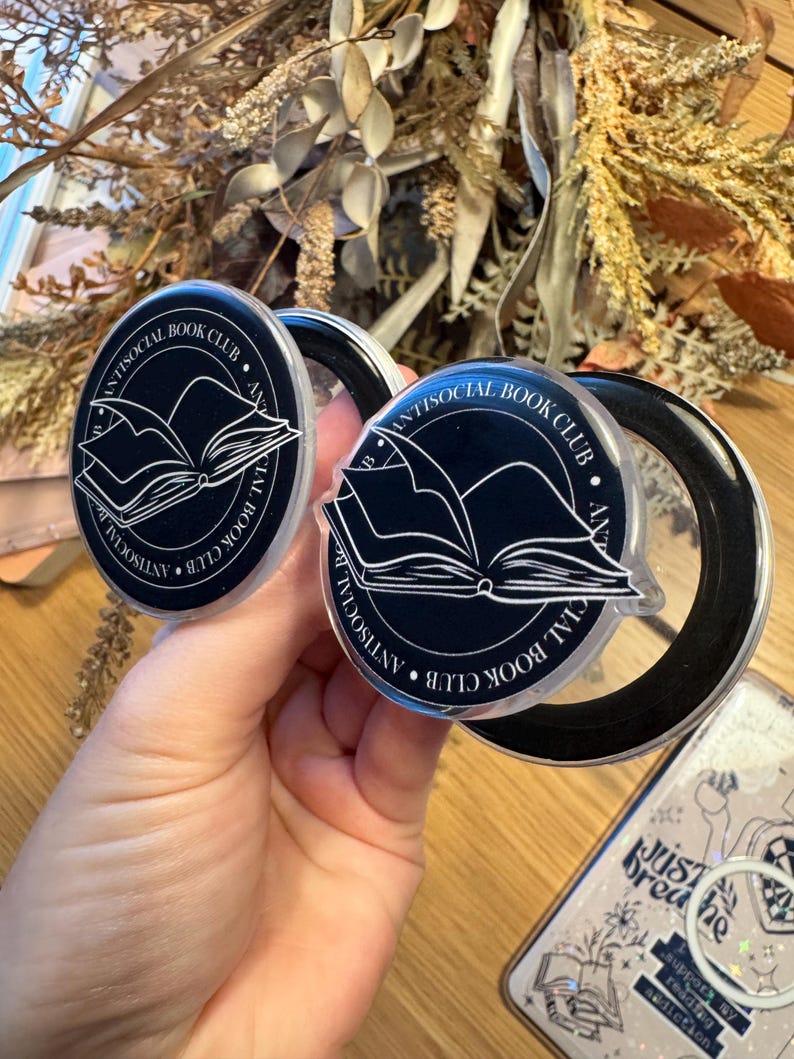 May include: Two black and clear phone grips with the text "ANTISOCIAL BOOK CLUB" and an open book graphic. The grips are round and held in front of a blurred background with dried flowers and a phone case.