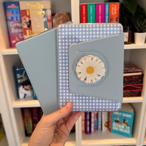 Peut inclure: Une housse de tablette bleu clair avec un motif vichy et un pop-socket en forme de marguerite. L'étui est ouvert, révélant l'écran de la tablette. L'étui est tenu devant une bibliothèque remplie de livres.
