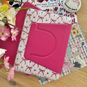 Könnte beinhalten: Eine leuchtend pinke E-Reader-Hülle mit klappbarem Ständer, verziert mit einem weiß-rosa Streifenmuster, kleinen roten Schleifen und Herzen. Die Hülle ist von verschiedenen dekorativen Aufklebern umgeben, darunter einer mit dem Text 'so many books, so little time' und ein runder, geisterförmiger Anstecker mit der Aufschrift 'BOOKISH GIRLIE'.