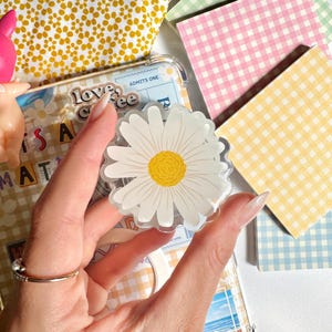 May include: A clear phone case with a beach scene and a daisy-shaped phone grip. The grip has a white flower with a yellow centre. Other items include a yellow floral pouch and pastel gingham notecards.