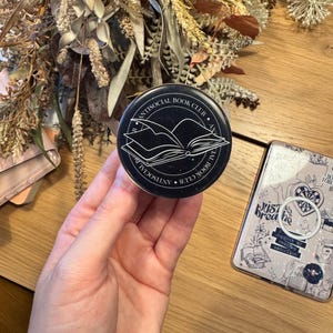 May include: A black and white round pop socket with the text "ANTISOCIAL BOOK CLUB" encircling an open book graphic. The pop socket is held in a hand, with a phone case and dried flowers in the background.