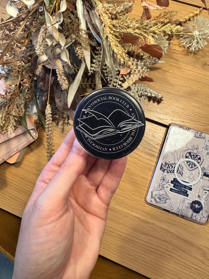 May include: A black and white round pop socket with the text "ANTISOCIAL BOOK CLUB" encircling an open book graphic. The pop socket is held in a hand, with a phone case and dried flowers in the background.