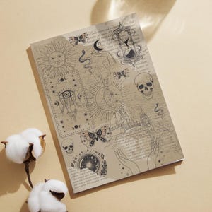 Op de afbeelding: Een beige notitieboek met een hemels ontwerp met een zon, maan, vlinders, schedels en een zandloper. Het notitieboek heeft een vintage esthetiek met gedrukte tekst en illustraties. Een glas water en katoenen bloemen staan op de achtergrond.