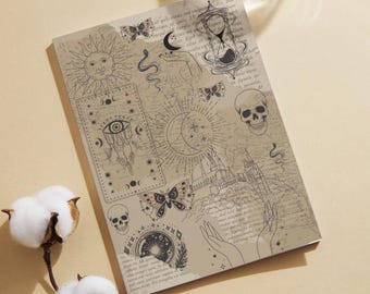 Op de afbeelding: Een beige notitieboek met een hemels ontwerp met een zon, maan, vlinders, schedels en een zandloper. Het notitieboek heeft een vintage esthetiek met gedrukte tekst en illustraties. Een glas water en katoenen bloemen staan op de achtergrond.