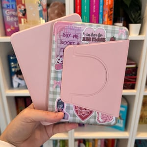 以下が含まれることがあります： ピンク色のタブレットケース。内蔵スタンド付きで、ステッカーで装飾されたタブレットを保持しています。「Buy Me Books」と「I'm here for Red Flags」というテキストが含まれています。背景には本棚が写っています。