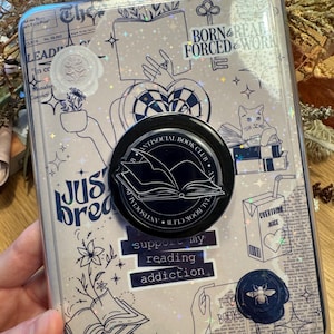 May include: A clear phone case with a book-themed design. The case features illustrations of books, hands, and a cat, along with text that reads "Born & Read Forced to Work" and "Support my reading addiction." A black pop socket with the words "Antisocial Book Club" is attached.