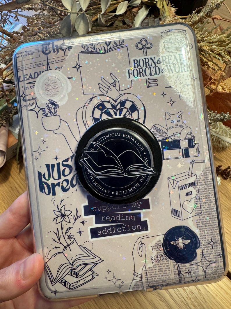 May include: A clear phone case with a book-themed design. The case features illustrations of books, hands, and a cat, along with text that reads "Born & Read Forced to Work" and "Support my reading addiction." A black pop socket with the words "Antisocial Book Club" is attached.