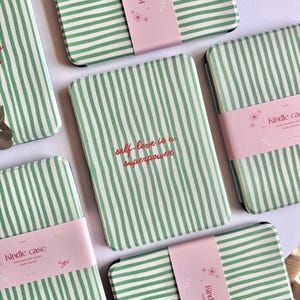 May include: Multiple rectangular e-reader cases featuring a green and white vertical striped pattern. One case prominently displays the red text "self-love is a superpower" in the center. Each case is wrapped in a light pink paper band labeled "kindle case".