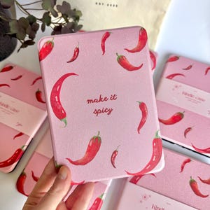 Puede incluir: Funda rosa para Kindle con un diseño de chiles rojos y la frase "make it spicy". Se ven varias fundas, junto con una bolsa de mano con el texto "lapazè".