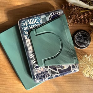 May include: A teal phone case with the words "MAGIC DRAGONS" and other stickers. A teal phone stand is attached to the case. A black and silver round object with an open book illustration is to the right.
