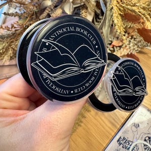 May include: Black and white phone grips with the text "Antisocial Book Club" encircling an open book illustration. The grips are round and have a pop-out feature for easy handling. A phone case with the text "Just Breathe" is also visible.
