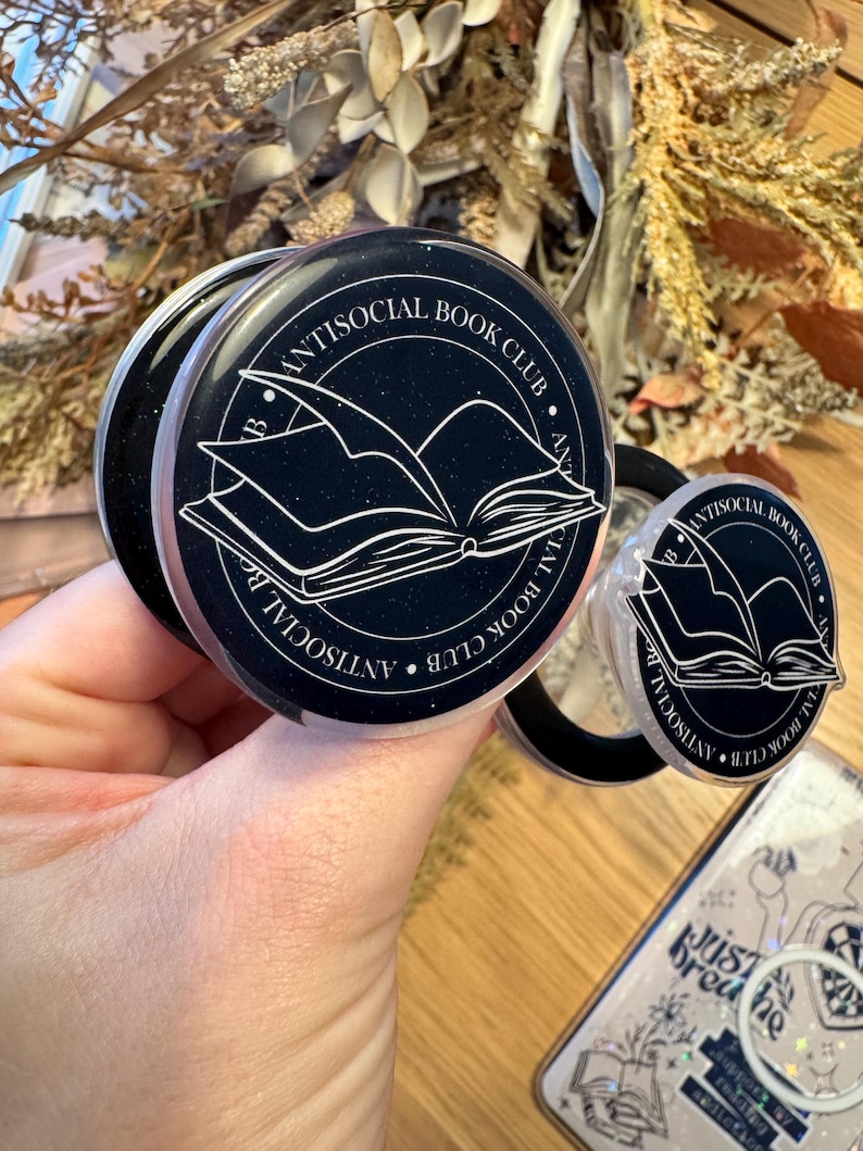 May include: Black and white phone grips with the text "Antisocial Book Club" encircling an open book illustration. The grips are round and have a pop-out feature for easy handling. A phone case with the text "Just Breathe" is also visible.