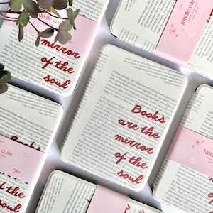 Puede incluir: Una colección de fundas para Kindle con una página de texto impresa de un libro. Cada funda muestra el texto rojo en cursiva "Books are the mirror of the soul" centrado en la página. Las fundas tienen un borde y detalles rosa claro.