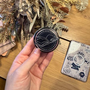May include: A black round pin with the text "ANTISOCIAL BOOK CLUB" encircling a graphic of an open book. The pin is held in a hand, with a phone case and dried flowers in the background.