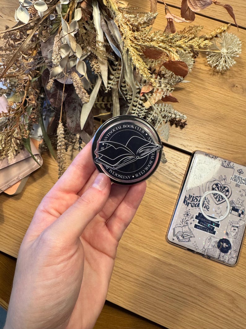 May include: A black round pin with the text "ANTISOCIAL BOOK CLUB" encircling a graphic of an open book. The pin is held in a hand, with a phone case and dried flowers in the background.