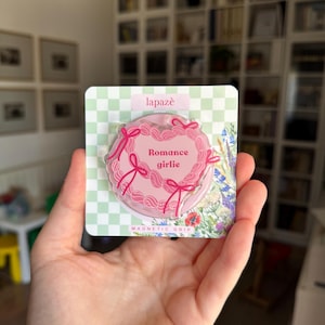 May include: A pink magnetic grip with the words "Romance girlie" in a heart shape, surrounded by pink bows and lace details. The grip is on a green and white checkered background, with the brand name "lapazè" at the top.