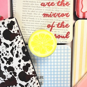 May include: A collection of e-reader cases featuring various patterns including a black and white cow print, a blue and white gingham check, yellow stripes, and a solid pink. A bright yellow lemon slice pop socket sits atop a book page.