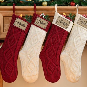 May include: Four cable-knit Christmas stockings hang from a wooden mantel. Two are red, and two are cream-colored. Each stocking has a personalized name tag. The mantel is decorated with greenery, ornaments, and a ribbon that reads "Merry Christmas."