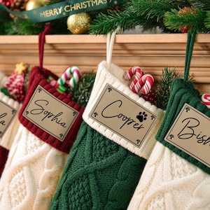 Puede incluir: Cuatro calcetines navideños personalizados en punto trenzado rojo, crema y verde. Cada calcetín tiene una etiqueta con un nombre. Los calcetines están decorados con bastones de caramelo y adornos navideños.