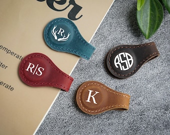 May include: Four leather golf ball markers in teal, burgundy, brown, and tan. Each marker features a white monogram or initial. The teal marker has a white antler design. The markers are stitched around the edges.