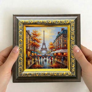 Pode incluir: Pintura emoldurada que retrata uma cena de rua chuvosa em Paris, com a Torre Eiffel ao fundo. A obra apresenta uma paisagem urbana detalhada com edifícios coloridos, árvores e pessoas com guarda-chuvas. A moldura é castanha com detalhes dourados.