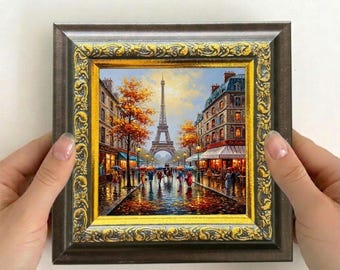 May include: A framed painting depicts a rainy street scene in Paris, with the Eiffel Tower in the background. The artwork features a detailed cityscape with colorful buildings, trees, and people with umbrellas. The frame is brown with gold accents.