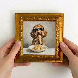 Op de afbeelding: Een ingelijst schilderij van een bruine hond met een zonnebril, die spaghetti eet uit een witte kom. Het schilderij heeft een gouden, sierlijke lijst. De hond is het middelpunt, met spaghettislierten uit zijn mond.