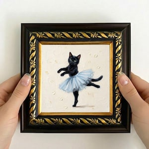 May include: A framed painting of a black cat in a light blue tutu, posed as a ballerina. The painting has a textured, off-white background with small, light-colored accents. The frame is dark brown with gold-colored decorative trim.