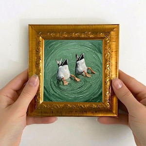 May include: A small, square painting in a gold ornate frame shows two ducks diving into green water. The ducks' white bodies and black tails are visible, with their orange feet sticking out. The painting is held by two hands.