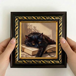 以下が含まれることがあります： 明るい琥珀色の目をした黒猫が、開いた本の上に頭と片足を乗せている様子を描いた正方形の額入り絵画。額縁はダークブラウンで、内側と外側の縁に金色の装飾的なディテールが施されています。