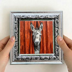 May include: A square oil painting featuring a grey donkey head emerging from behind thick, textured, vertical orange curtains. The artwork is encased in a decorative, ornate silver-toned frame with intricate embossed scrollwork patterns.