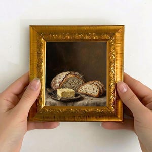 May include: A framed miniature painting of bread and butter. The painting depicts a loaf of bread cut in half, sliced bread, and a square of butter on a small plate. The frame is gold with ornate detailing.