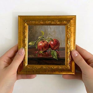 May include: A small, framed painting featuring two ripe pomegranates with green leaves and small red seeds. The artwork is set against a dark wooden surface and a neutral background. The frame is ornate and gold-colored.