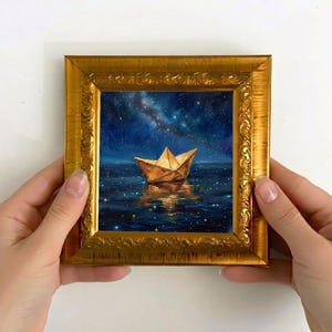 May include: A gold-framed painting shows a paper boat on water beneath a starry night sky. The artwork features a gold ornate frame. The painting's colours include deep blues, gold, and touches of white.