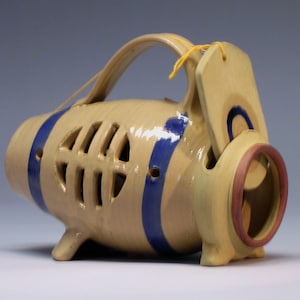 May include: A ceramic, beige-colored, hand-painted, decorative, hanging planter with a blue and white design. The planter has a handle and a small, round opening on the side. The planter is hanging from a yellow string.