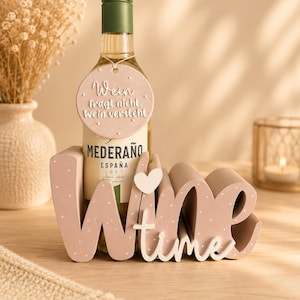 Könnte beinhalten: Eine Flasche Mederaño-Wein mit einem runden, hellrosa Anhänger mit weißen Punkten und dem Text "Wein fragt nicht. Wein versteht." Die Flasche steht in einem hellrosa Holzhalter in Form des Wortes "Wine" mit einem kleinen Herz.