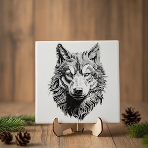 May include: A square, white ceramic tile featuring a detailed black and white illustration of a wolf's head. The tile is displayed on a small wooden stand, with pine needles and pine cones in the background.