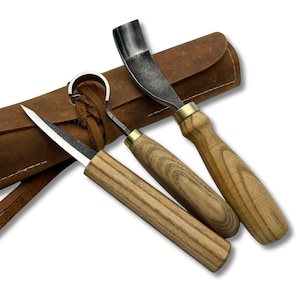 May include: A set of three wood carving tools with wooden handles and metal blades, alongside a brown leather sheath. The tools include a knife, a hook knife, and a curved gouge. The sheath has a loop closure.