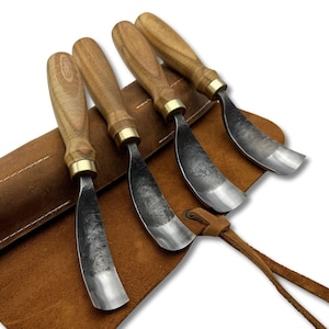 May include: A set of four wood carving gouges with wooden handles and metal blades, displayed in a brown leather tool roll. The gouges have brass ferrules and are arranged at an angle, showcasing their curved blades.