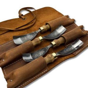 May include: A set of four wood carving gouges with wooden handles and metal blades, nestled in individual leather sheaths. The tools are arranged in a brown leather roll-up case with a tie closure. The gouges have brass ferrules.