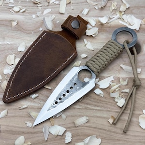 May include: A handmade throwing knife with a sharp, silver blade and a tan-colored wrapped handle. The knife features a ring at the end of the handle and is accompanied by a brown leather sheath. The knife is approximately 8 inches long.