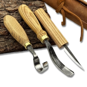 May include: A set of three wood carving tools with wooden handles and metal blades. The handles are light-colored wood with brass accents. One tool has a curved blade, another a gouge, and the third a straight blade. A brown leather sheath is visible in the background.