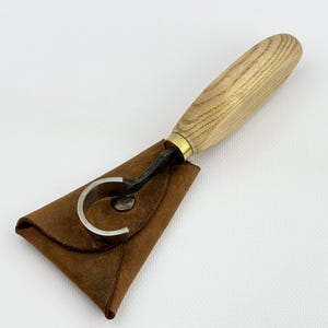 May include: A wood carving knife with a curved blade and a wooden handle. The knife is in a brown leather pouch with a snap closure. The handle is light-colored wood with a brass ferrule.