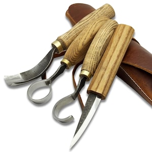 Puede incluir: Un juego de cuatro herramientas para tallar madera con mangos de madera y hojas de metal. Las herramientas están dispuestas en fila, con una funda de cuero en el fondo. Los mangos son de color marrón claro con un patrón de vetas de madera y las hojas son plateadas.